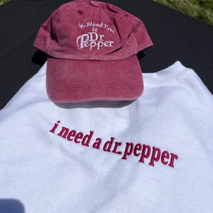 May include: A white t-shirt with the text "i need a dr pepper" embroidered in pink. A red baseball cap with the text "My Blood Type is Dr Pepper" embroidered in white is on top of the shirt.