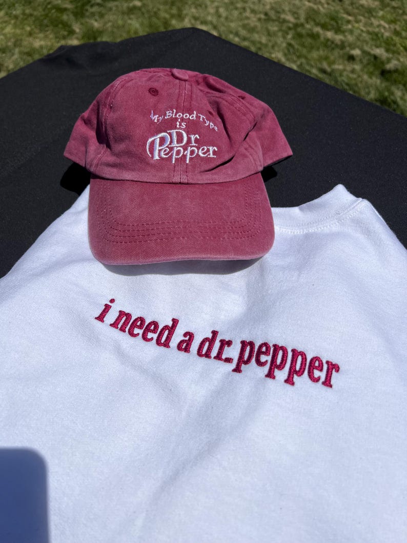 May include: A white t-shirt with the text "i need a dr pepper" embroidered in pink. A red baseball cap with the text "My Blood Type is Dr Pepper" embroidered in white is on top of the shirt.
