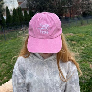 May include: A pink baseball cap with the text "Faith over Fear" embroidered in white. The cap has a white arrow pointing to the right.