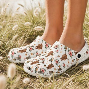 May include: White clogs with a repeating pattern of brown Highland cows and pink and yellow flowers. The clogs have a strap and are worn in a grassy field.