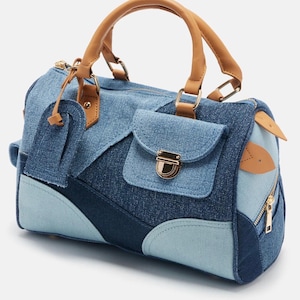 Puede incluir: Bolso de mano vaquero con diseño patchwork en varios tonos de azul. El bolso tiene un asa superior y un pequeño bolsillo delantero con un cierre dorado. Las asas y los ribetes son de color marrón claro. Una cremallera es visible en el lateral.