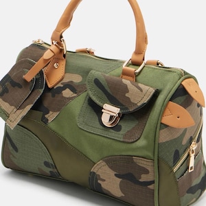 May include: A green and camouflage print handbag with tan leather handles and accents. The bag features a top handle, a front pocket with a gold clasp, and a side zipper. The camouflage pattern includes shades of brown, green, and black.