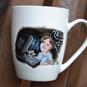 Op de afbeelding: Witte keramische mok met een handgeschilderd ontwerp van een persoon die aan een computer werkt. De persoon heeft bruin haar en draagt een lichtblauw shirt. De achtergrond is zwart met witte wolken.