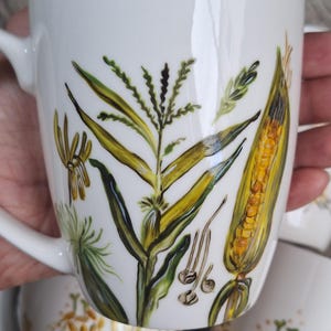 Op de afbeelding: Witte keramische mok met een handgeschilderd ontwerp van maïsplanten, bladeren en een maïskolf in geel, groen en bruin. De mok heeft een gebogen handvat en een gladde, glanzende afwerking. Het ontwerp is gedetailleerd en botanisch.