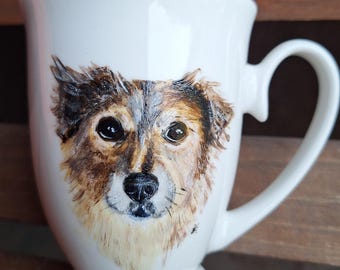 May include: White ceramic mug with a hand-painted portrait of a dog. The dog has brown, black, and white fur and is looking directly at the viewer.