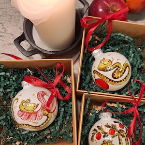 Puede incluir: Tres adornos navideños blancos con ilustraciones pintadas a mano de serpientes verdes sobre fondo blanco. Los adornos están en cajas individuales con papel verde triturado.