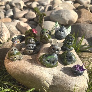 May include: A collection of small, whimsical stone sculptures, each uniquely designed with moss, crystals, and miniature details. The sculptures are arranged on a large, light-colored rock, surrounded by other rocks and greenery.