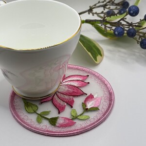 Può includere: Un sottobicchiere ricamato rosa con un motivo floreale. Il sottobicchiere è realizzato in un tessuto delicato e trasparente e presenta un fiore rosa con foglie verdi.