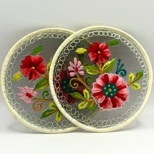 May include: Two round embroidered patches with a floral design. The patches are white with a delicate lace trim and feature pink, red, and green flowers.