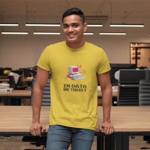 Puede incluir: Una camiseta amarilla con el texto "IN DATA WE TRUST" y un gráfico de una computadora retro. La persona lleva vaqueros azules. La imagen está tomada en un entorno de oficina.