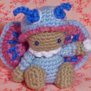 May include: A crocheted blue and pink butterfly with a white face and blue eyes. The butterfly is wearing a blue and white crocheted hat.