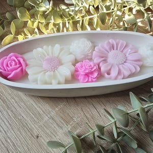 Può includere: Una ciotola ovale in cemento piena di candele a forma di fiore. Le candele sono in tonalità di rosa e bianco, tra cui rose, margherite e altri disegni floreali. La ciotola è appoggiata su una superficie di legno, con fogliame verde sullo sfondo.