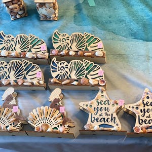 May include: Wooden beach-themed decorations featuring seashell accents. Includes laser-cut seashell designs, mermaid figures, and starfish with the text "sea you at the beach". The items are displayed on a blue surface.