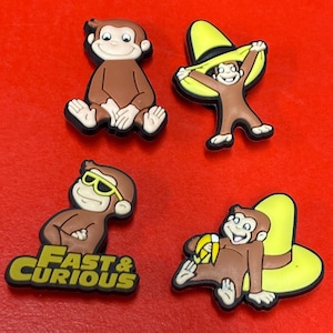 May include: Four brown monkey-themed shoe charms on a red surface. One charm shows a monkey sitting, another holding a hat, a third wearing sunglasses with the text "Fast & Curious", and the last holding a banana.