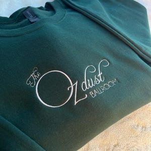 Puede incluir: Una sudadera verde oscuro con texto bordado en blanco que dice "The Oz dust Ballroom".