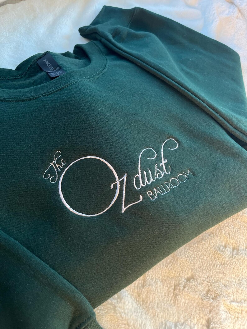 Puede incluir: Una sudadera verde oscuro con texto bordado en blanco que dice "The Oz dust Ballroom".