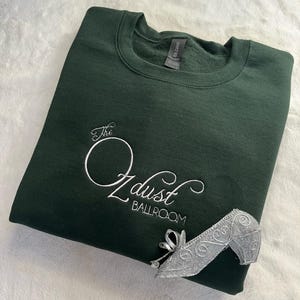 Puede incluir: Una sudadera verde oscuro con el texto "The Oz dust Ballroom" bordado en blanco. Un zapato de plata est&aacute; sobre la sudadera.