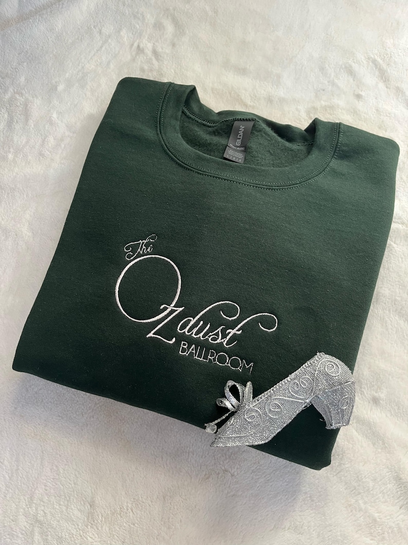 Puede incluir: Una sudadera verde oscuro con el texto "The Oz dust Ballroom" bordado en blanco. Un zapato de plata est&aacute; sobre la sudadera.