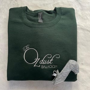 Puede incluir: Una sudadera verde oscuro con el texto "The Ozdust Ballroom" bordado en blanco en la parte delantera. Un zapato de charol plateado est&aacute; sobre la sudadera.