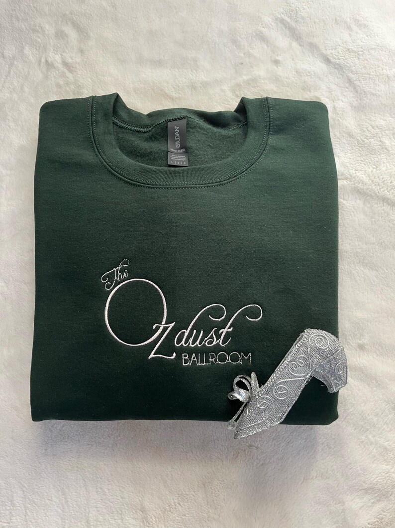 Puede incluir: Una sudadera verde oscuro con el texto "The Ozdust Ballroom" bordado en blanco en la parte delantera. Un zapato de charol plateado est&aacute; sobre la sudadera.