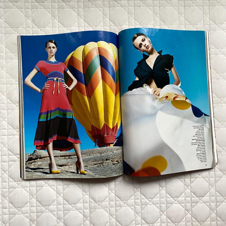 May include: Two women wearing colorful clothing pose in front of a hot air balloon. The woman on the left is wearing a red, black, and white dress. The woman on the right is wearing a black top and a white skirt with yellow and red polka dots. The background is a bright blue sky.