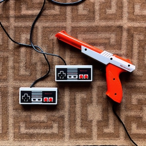 May include: Two white Nintendo controllers with red buttons and a white and orange Nintendo Zapper gun. The controllers and gun are on a brown patterned surface.