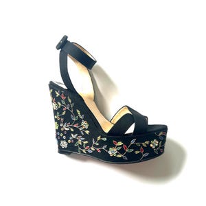 May include: A black wedge sandal featuring an ankle strap with a buckle closure and an open toe design. The exterior of the wedge heel and platform is decorated with an intricate, colorful floral embroidery pattern accented with small, sparkling crystals.