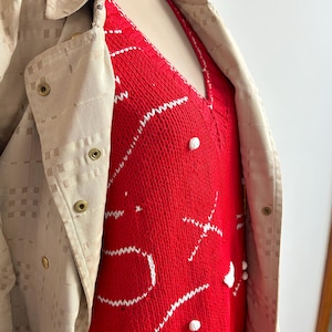 May include: A red knit sweater with white embroidered details and small white pom-poms. A tan jacket with a grid pattern and snap closures is partially visible on the left side of the image.