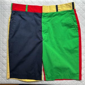 May include: Colour-blocked shorts in navy blue and green, with red and yellow accents. The shorts have a classic design with pockets and a button closure. The waistband is red and yellow, with a green stripe on the side.