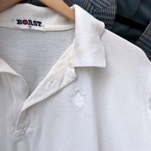 May include: White polo shirt with a collared neckline and a three-button placket. The shirt features a small embroidered leaf design on the chest and the brand name "BOAST" on the collar.