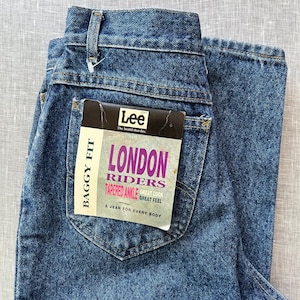Pode incluir: Close-up de calças de ganga azuis com uma etiqueta da marca "Lee" no bolso traseiro. A etiqueta diz "LONDON RIDERS" com o texto "TAPERED ANKLE" e "BAGGY FIT". As calças de ganga têm um design clássico de cinco bolsos.
