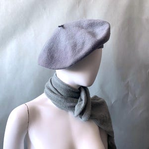 May include: A gray beret with a slightly darker shade of gray on the top. The beret is on a white mannequin head with a gray scarf around the neck.