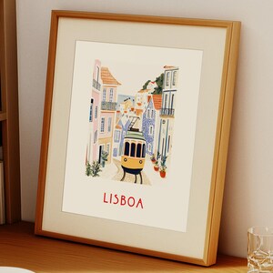 May include: A framed illustration of a yellow tram traveling up a hill in Lisbon, Portugal. The buildings are colorful and the street is cobblestone. The text "LISBOA" is printed in red at the bottom of the image.