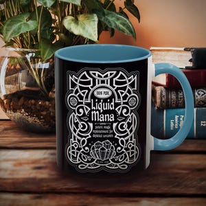 May include: Black and white ceramic mug with a blue interior and handle. The mug features the text "100% Pure Liquid Mana" in a decorative design. The mug is 11 fl oz.