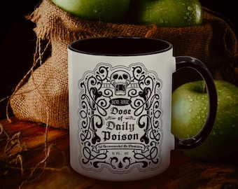 Puede incluir: Una taza de cerámica blanca con interior y asa negros, con un diseño decorativo en blanco y negro con una calavera y el texto "Dose of Daily Poison". La taza es de 325 ml y está junto a manzanas verdes y un saco de yute.