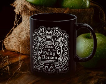 Puede incluir: Taza de cerámica negra con el texto "SEMI-TOXIC Dose of Daily Poison" en un diseño blanco decorativo. La taza contiene 325 ml de líquido. Hay manzanas verdes en el fondo.