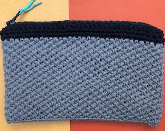 Peut inclure: Une pochette crochetée à la main dans les tons de gris et de bleu marine. La pochette présente un motif de point texturé et une fermeture éclair avec une tirette turquoise. Le fond est divisé en sections jaune et rouge.