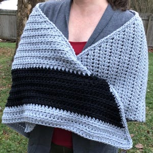 May include: A handmade crochet shawl in a triangular shape. The shawl features a light gray color with a thick black stripe across the center. The shawl is worn over a gray hooded shirt and a red top.