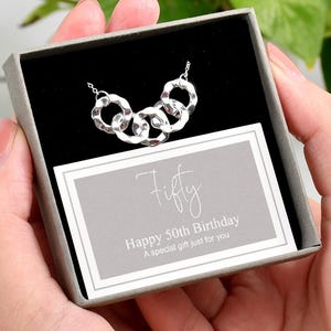 Könnte beinhalten: Eine silberne Halskette mit fünf ineinandergreifenden Kreisen wird in einer Geschenkbox präsentiert. Die Box enthält eine Karte mit der Aufschrift "Fifty" und "Happy 50th Birthday". Der Hintergrund zeigt grünes Laub.