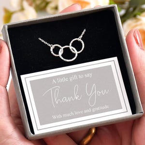 May include: A silver necklace with three linked circles is presented in a grey gift box. The box contains a card that reads "A little gift to say Thank You with much love and gratitude."