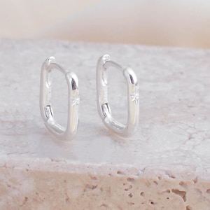 May include: Two silver rectangular hoop earrings. They are presented on a light-coloured, textured surface, set against a soft, cream-coloured backdrop. The earrings have a simple, contemporary design.