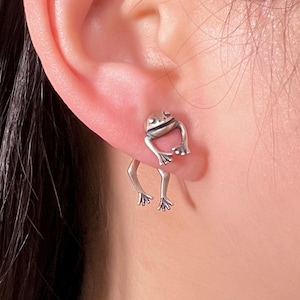 Könnte beinhalten: Silberne Frosch-Ohrringe mit einem hängenden Design. Der Frosch sitzt auf einer kleinen Plattform mit seinen Beinen, die nach unten hängen.