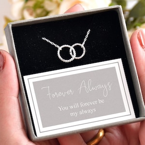 Può includere: Una collana d'argento con due cerchi intrecciati, ognuno con piccoli ciottoli trasparenti. La collana è presentata in una scatola grigia con un biglietto bianco su cui è scritto "Forever Always. You will forever be my always."