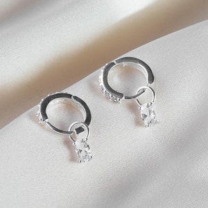 May include: A pair of silver hoop earrings with small clear stones set around the hoop. Each earring has a small, dangling, clear, rectangular stone. The earrings are displayed on a white, silky fabric.