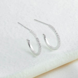 Puede incluir: Un par de pendientes de aro de plata con una fila de pequeñas piedras de circonita cúbica transparentes alrededor del borde.