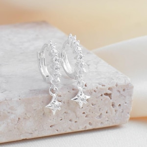 May include: A pair of silver hoop earrings with a star-shaped charm dangling from each hoop. The earrings are set with small, clear stones.