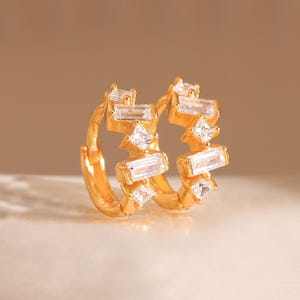 May include: A pair of gold-coloured hoop earrings. Each earring features multiple clear, rectangular and square-shaped gemstones. The earrings are displayed on a light-coloured surface against a soft, neutral background.
