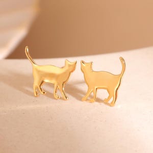 May include: A pair of gold-colored cat-shaped earrings. The earrings are in the shape of a cat silhouette, with a tail and four legs. The earrings are on a light-colored surface.