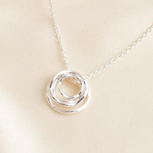 Può includere: Una collana d'argento con tre cerchi intrecciati. Il ciondolo pende da una catena sottile, offrendo un design minimalista ed elegante. I cerchi hanno una finitura strutturata, aggiungendo interesse visivo al gioiello.