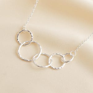 May include: A silver necklace featuring five interconnected textured rings. The rings are of varying sizes and are linked together in a cascading design. The necklace has a delicate chain and is displayed on a cream-colored fabric.